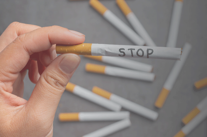 Foto horizontal: mão segura um cigarro com a palavra “STOP”, enquanto outros cigarros aparecem espalhados ao fundo.