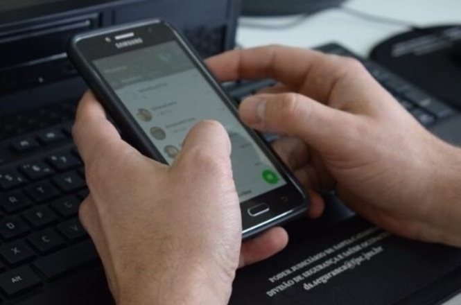 Foto horizontal: mãos seguram um celular durante o uso do aparelho, com teclado de computador ao fundo.