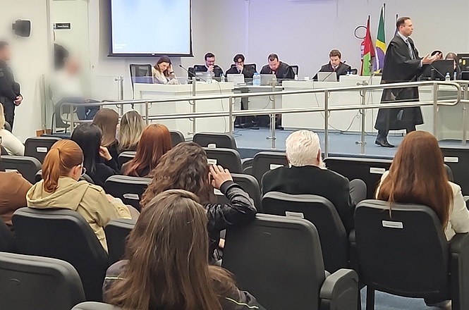 Foto horizontal: sessão de julgamento em auditório, com magistrados à mesa e público acompanhando a atividade.