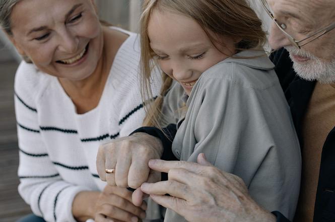 Foto horizontal: menina aparece sorrindo entre uma mulher e um homem idosos, em momento de carinho e convivência familiar.