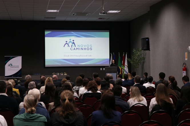 Foto horizontal: público acompanha apresentação do projeto Novos Caminhos em auditório, com palestrante ao lado do telão.