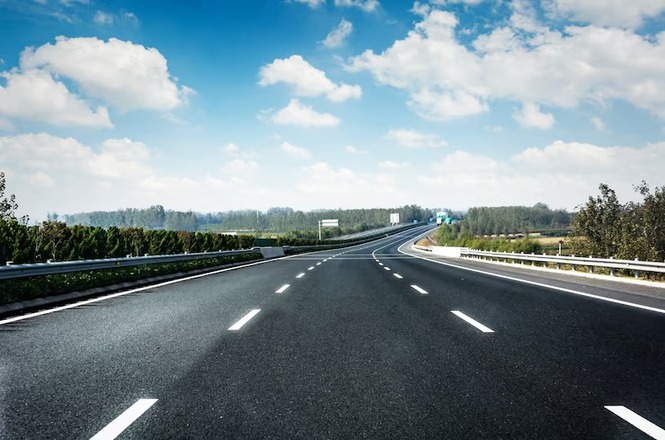 Foto horizontal: rodovia asfaltada de pista dupla, com faixas de sinalização brancas, cercada por vegetação sob céu azul com nuvens.
