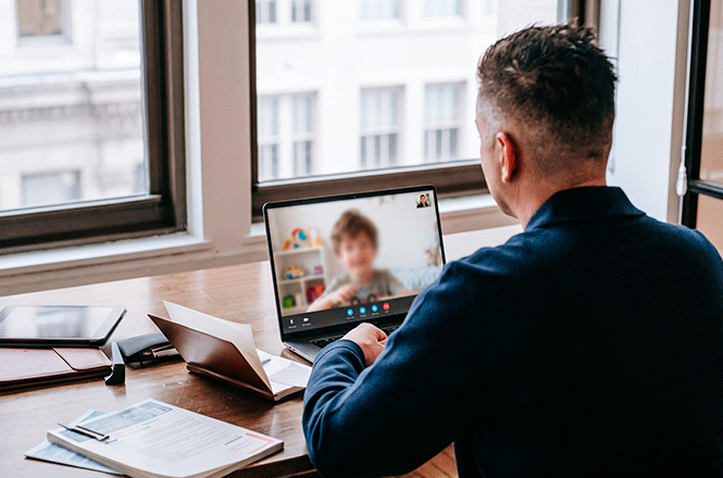 Foto horizontal: homem participa de videoconferência pelo notebook, sentado à mesa com documentos e outros materiais de trabalho.