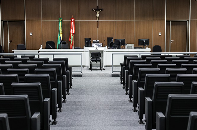 Foto horizontal: plenário vazio, com fileiras de cadeiras, mesa de julgamento, bandeiras do Brasil e de Santa Catarina e crucifixo ao fundo.