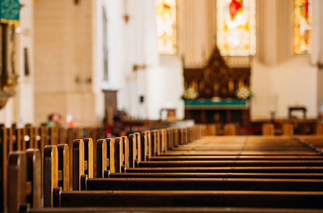 Foto horizontal: interior de igreja com fileiras de bancos de madeira voltadas para o altar, iluminado por vitrais ao fundo.