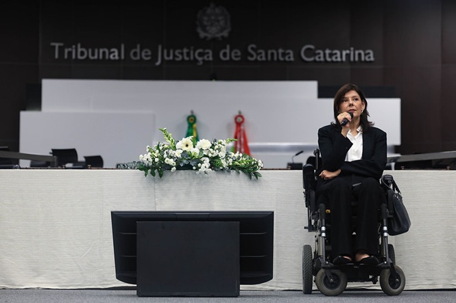 Foto horizontal: jornalista e escritora Flávia Cintra fala ao microfone, em cadeira de rodas, no plenário do TJSC.