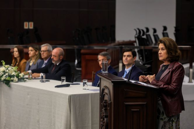 Fotografia horizontal, em ambiente de auditório. Em primeiro plano, à direita, a desembargadora Vera Lúcia Copetti, diretora-executiva da Academia Judicial, está em pé ao lado de um púlpito de madeira, onde faz seu pronunciamento. Ela veste blazer em tom vinho sobre blusa escura. Ao fundo, à esquerda, há uma mesa de autoridades com várias pessoas sentadas, decoração floral e microfones. O ambiente possui paredes e mobiliário em tons escuros, com o plenário ao fundo.
