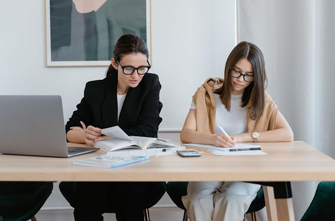 Foto horizontal: duas mulheres analisam documentos e fazem anotações em uma mesa, com notebook e calculadora ao lado.