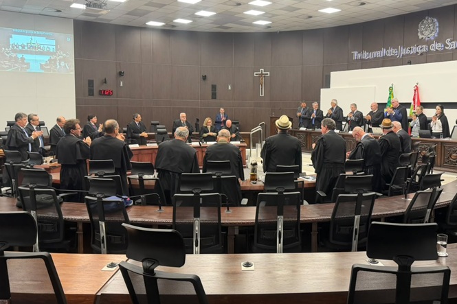 Foto horizontal: magistrados em pé participam de sessão no Tribunal Pleno do TJSC, com crucifixo e bandeiras ao fundo.