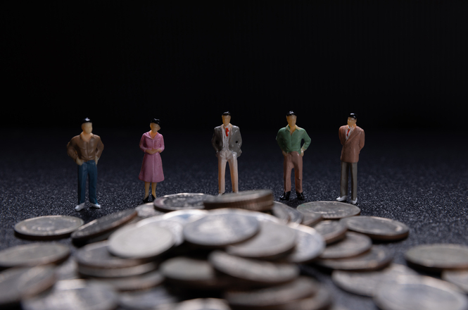 Foto horizontal: miniaturas de pessoas aparecem atrás de uma pilha de moedas, em representação de questões financeiras.