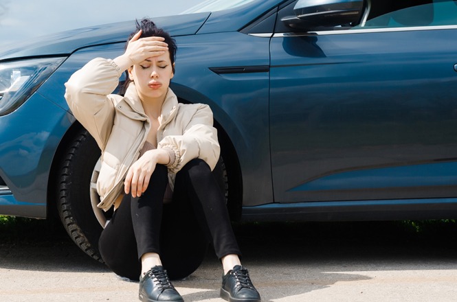 Foto horizontal: mulher sentada no chão, ao lado de um carro azul, leva a mão à testa com expressão de preocupação.