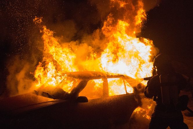 Foto horizontal: veículo é consumido por grandes chamas enquanto bombeiro atua no combate ao incêndio.