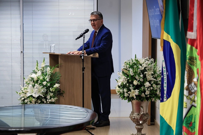 Foto horizontal: juiz Augusto Cesar Allet Aguiar discursa ao microfone em púlpito, com bandeiras e arranjos de flores ao lado.