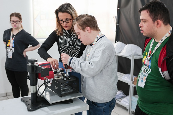 Foto horizontal: jovens participam de atividade prática com equipamento de personalização, acompanhados por instrutora.