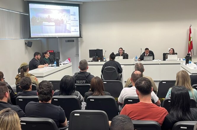 Foto horizontal: público acompanha sessão em sala de audiências, com magistrados à frente e projeção em tela.