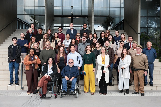 Foto horizontal: grupo de participantes posa para foto em frente a edifício, incluindo pessoas com deficiência e mobilidade reduzida.