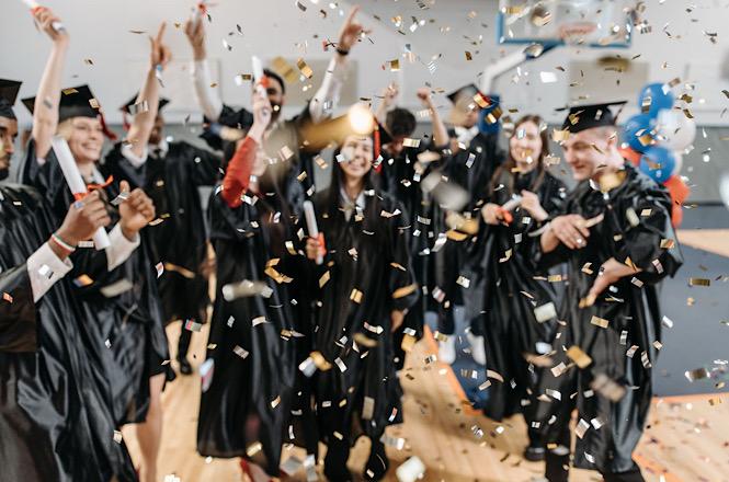 Foto horizontal: grupo de formandos celebra conclusão de curso com becas, capelos e chuva de confetes.