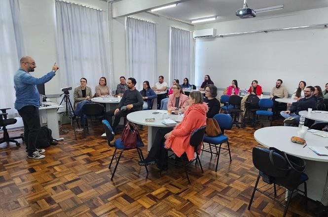 Foto horizontal: palestrante conduz atividade com grupo de participantes reunido em sala de aula, distribuído em mesas.
