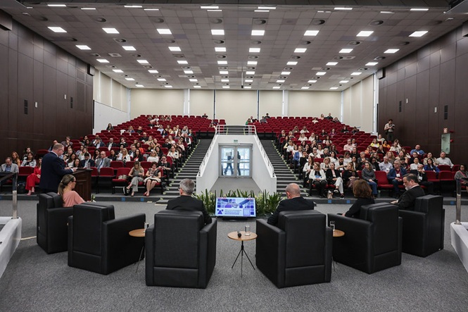 Foto horizontal: público acompanha evento em auditório, enquanto autoridades participam de atividade no espaço central.