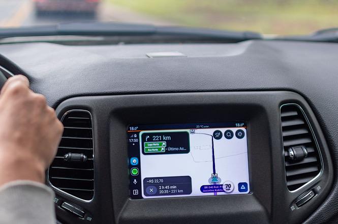 Foto horizontal: motorista conduz veículo enquanto painel multimídia exibe rota de navegação por GPS.