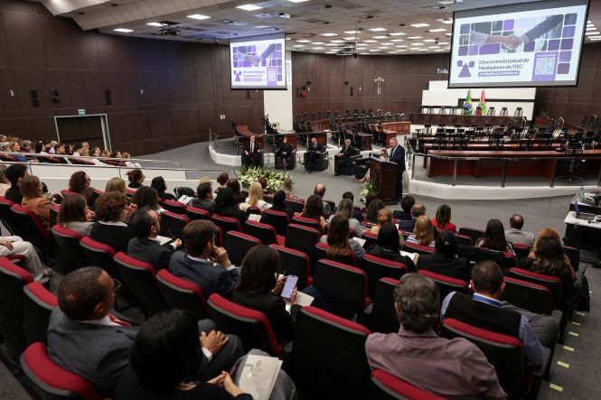 Fotografia em plano aberto de um auditório de grande porte durante um evento institucional. Dezenas de participantes estão sentados em poltronas vermelhas, voltados para a parte frontal da sala. No palco, há uma mesa de autoridades e uma pessoa discursando em um púlpito. Ao fundo, duas grandes telas exibem a identidade visual do evento, com a inscrição “4º Encontro Estadual de Mediadores e Mediadoras de Justiça”. O ambiente possui paredes em madeira escura, iluminação artificial e bandeiras posicionadas ao lado do palco.