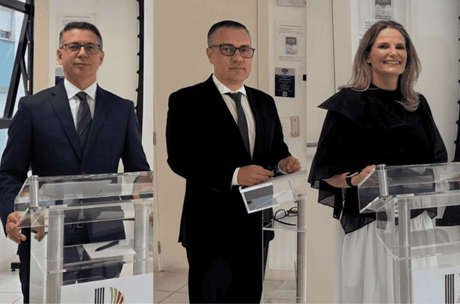 Foto horizontal: duas autoridades de terno escuro e mulher de preto e branco posam junto a púlpitos transparentes.