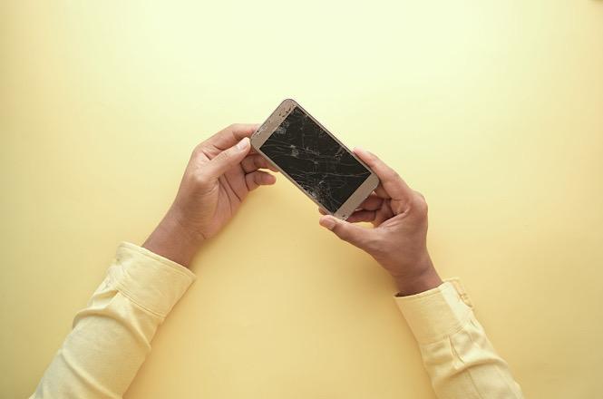 Foto horizontal: pessoa segura celular com a tela quebrada, em representação de dano ao aparelho.