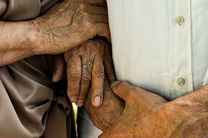 Foto horizontal: mãos de duas pessoas idosas se entrelaçam em gesto de carinho, cuidado e companheirismo.