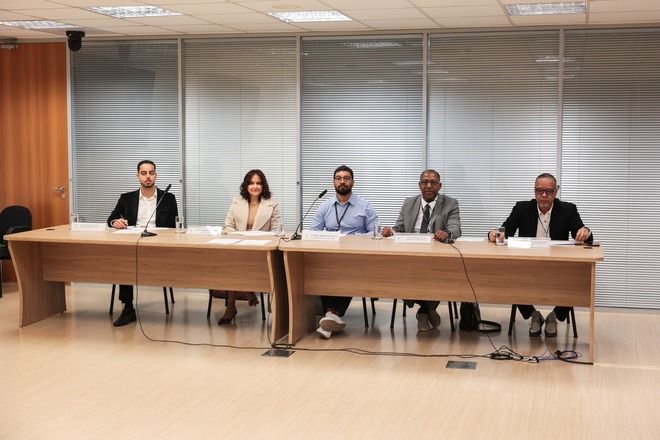 Foto horizontal: cinco participantes integram mesa de debates, com microfones e documentos dispostos à frente.