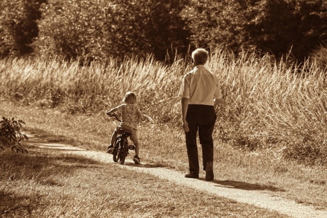 Foto horizontal: criança pedala por caminho rural enquanto homem caminha ao seu lado, em cena de convivência familiar.