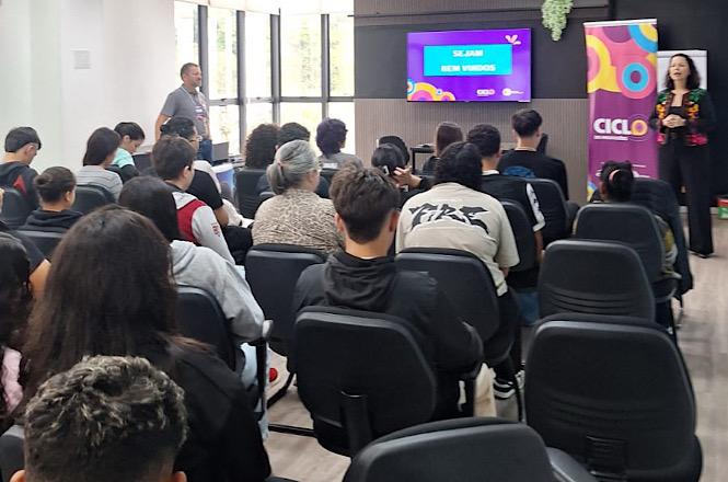 Foto horizontal: palestrante de preto fala a jovens sentados em auditório, com tela e banner coloridos ao fundo.