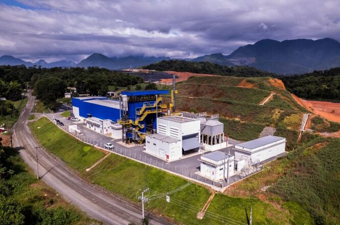 Foto horizontal: complexo industrial com estruturas azuis e brancas, cercado por vegetação, estrada e montanhas.