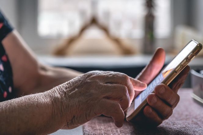 Foto horizontal: pessoa idosa segura celular e toca a tela com o dedo, em ambiente doméstico.