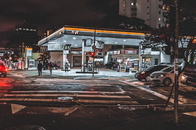 Foto horizontal: posto de combustíveis iluminado à noite, com bombas de abastecimento e veículos estacionados.