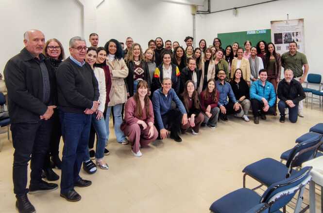Foto horizontal: grupo numeroso posa reunido em sala, com participantes em pé e agachados diante de cadeiras.
