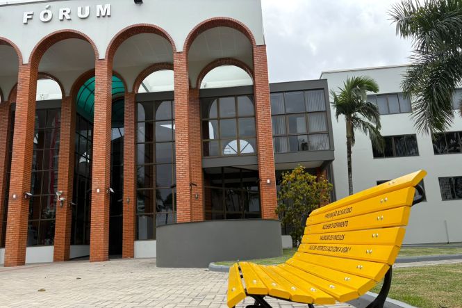 Foto horizontal: banco amarelo em primeiro plano diante do Fórum de Tubarão, com fachada de arcos e palmeiras.
