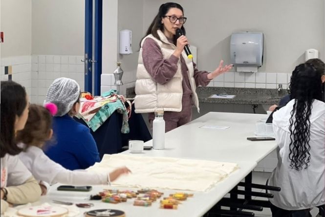 Foto horizontal: mulher de colete bege fala ao microfone para grupo reunido em sala, com materiais sobre a mesa.