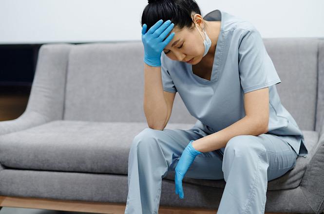 Anexo do usuário Foto horizontal: profissional de saúde de uniforme cinza e luvas azuis descansa sentada, com a mão sobre a testa.