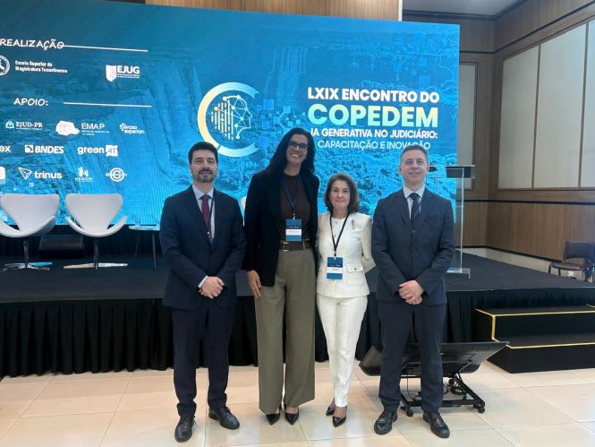 Da esquerda para a direita, Gustavo Heil, usando terno azul-marinho, camisa branca e gravata bordô; a juíza Fernanda Nunes Pereira, vestindo blazer preto, blusa escura, calça bege e crachá de identificação; a desembargadora Vera Copetti, com conjunto social branco composto por blazer e calça, além de crachá; e o juiz Jefferson Zanini, usando terno cinza-escuro, camisa clara e gravata azul. Os quatro estão posicionados em frente ao palco do LXIX Encontro do Copedem. Ao fundo, um painel azul exibe o tema do evento, “IA Generativa no Judiciário: Capacitação e Inovação”, além de logotipos de instituições parceiras e apoiadoras. Também são visíveis poltronas brancas sobre o palco e parte da estrutura do auditório.