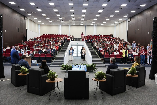 Foto horizontal: público ocupa auditório de poltronas vermelhas durante evento, diante de palco com cadeiras pretas.