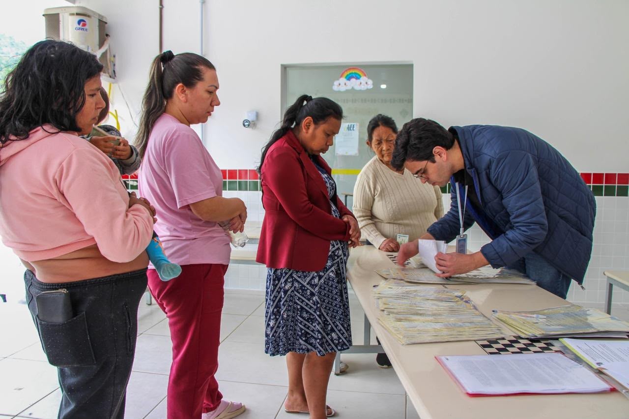 Foto colorida: pessoas em fila diante de mesa de atendimento, sala comunitária interna, ação social, conferindo documentos em rosa, azul e branco