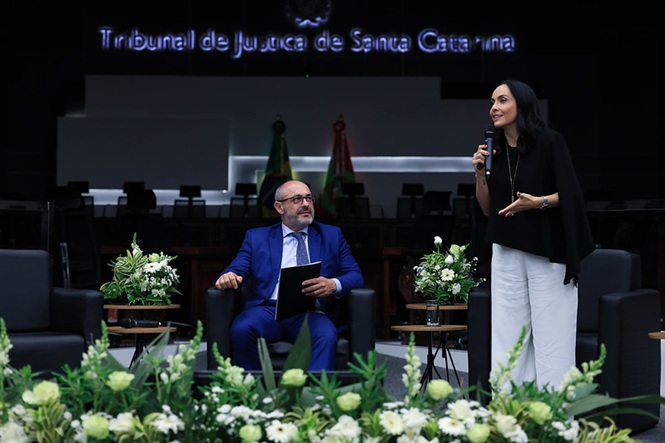 Des. André Dacol, de terno azul, observa a jornalista Izabella Camargo, de preto e branco, falando ao microfoneplenário do TJSC, evento institucional, apresentando tema em preto, azul e branco