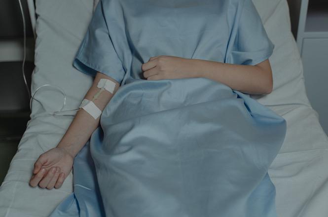 Foto colorida: paciente deitado em leito hospitalar, quarto clínico interno, atendimento de saúde, recebendo medicação em azul, branco e bege