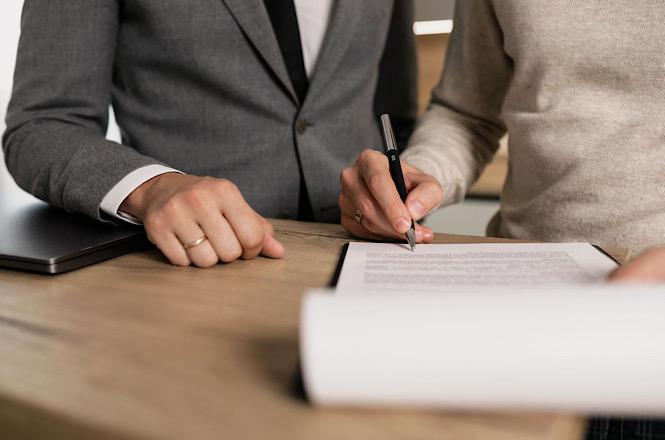 Horizontal: Pessoa assina documento sobre mesa de escritório, em ambiente corporativo com notebook e papéis ao redor.