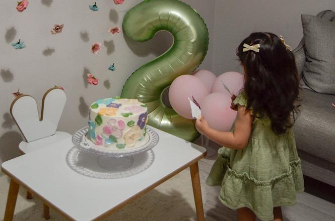 Horizontal: Criança ao lado de bolo decorado e balões em sala de festa, com sofá, mesa branca e parede enfeitada.