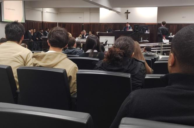 Foto horizontal: público acompanha sessão em tribunal, com magistrado à frente e crucifixo na parede.