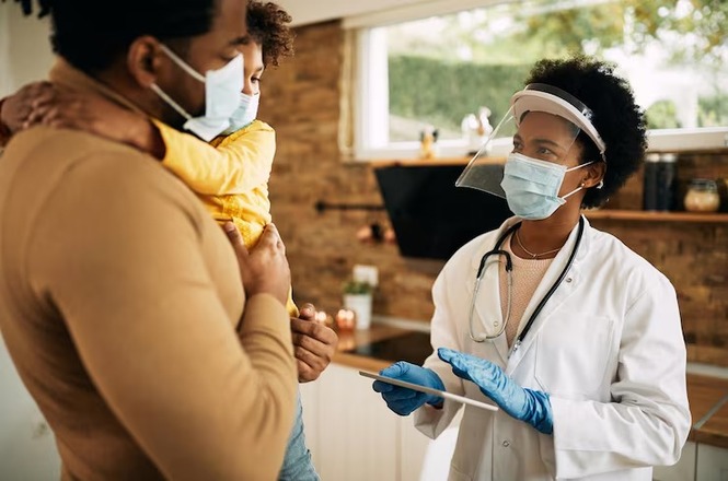 Foto horizontal: médica de branco, máscara e protetor facial orienta homem de marrom que segura criança de amarelo.