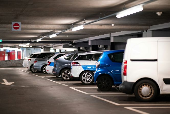 Foto horizontal: veículos brancos, cinza e azul estão estacionados lado a lado em garagem coberta e iluminada.