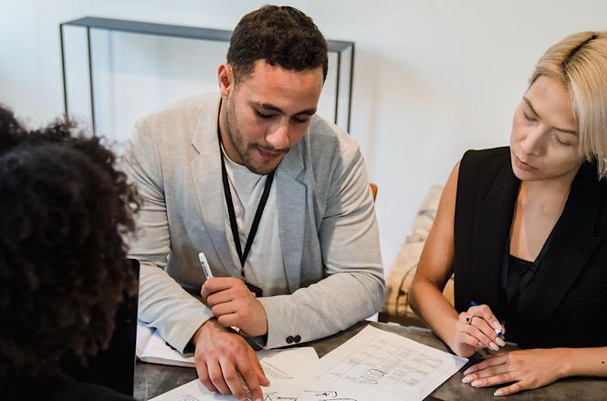 Foto horizontal: homem de blazer cinza e mulher de preto analisam e fazem anotações em documento sobre a mesa.