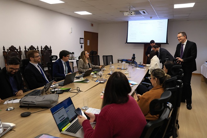 Foto horizontal: equipe trabalha com notebooks em reunião, enquanto servidor de terno e gravata roxa orienta o grupo.